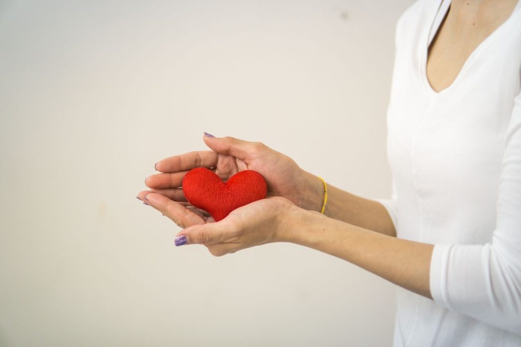 Unrecognizable lady wearing white outfit standing on white background with decorative small red heart in hands in light room inside